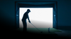 Silhouette of a golfer practicing indoors with a bright light source behind him.